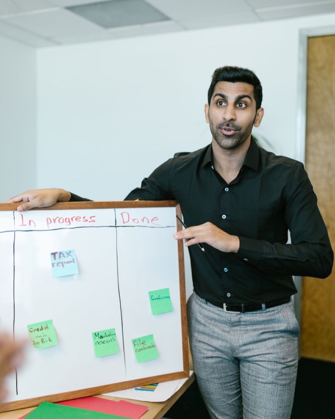 A young adult male presents project progress using a whiteboard in a modern office setting.