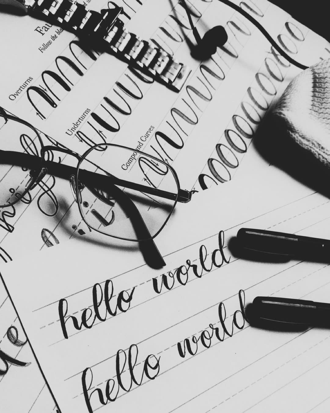 Monochrome image of calligraphy practice with eyeglasses and pens on sheets of paper.
