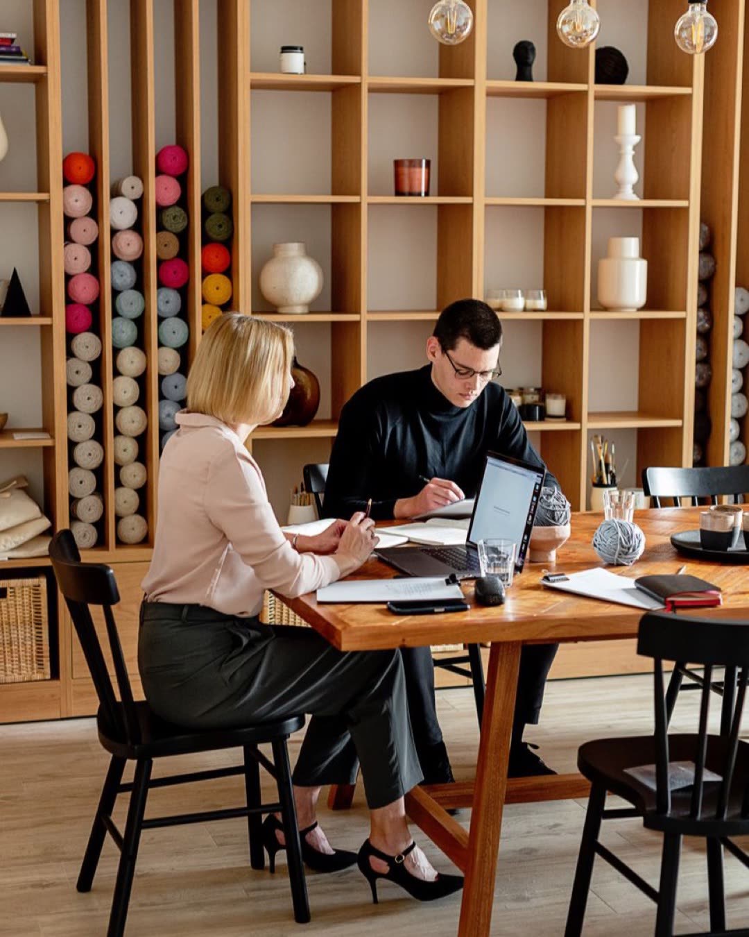 Two colleagues working together at a wooden desk in a modern office with yarn display in background.