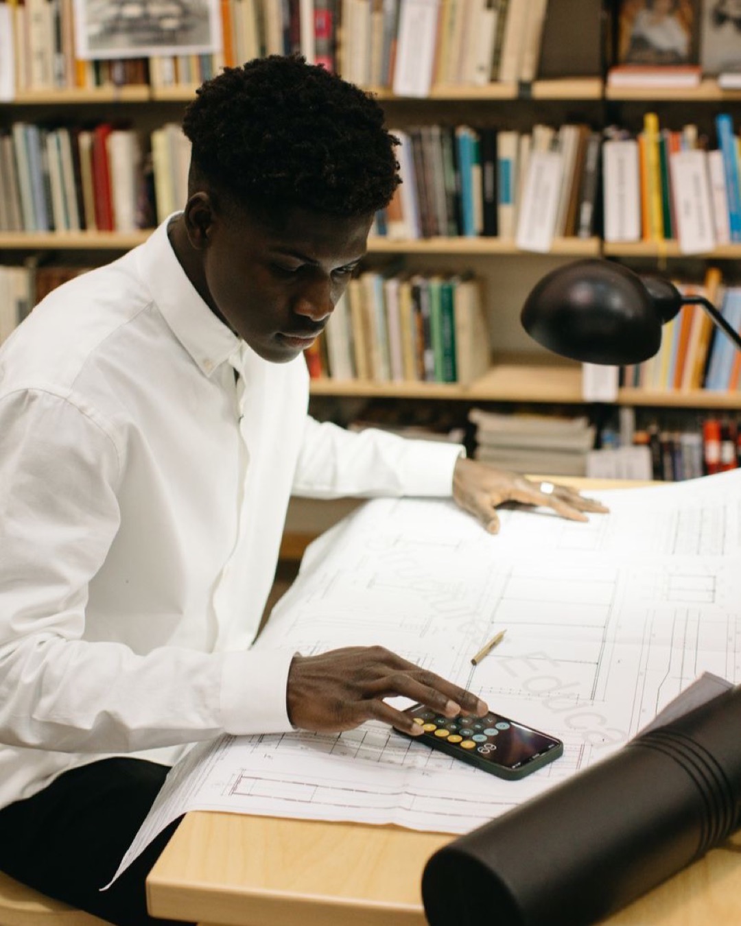Architect in library calculating figures on large blueprints at desk.