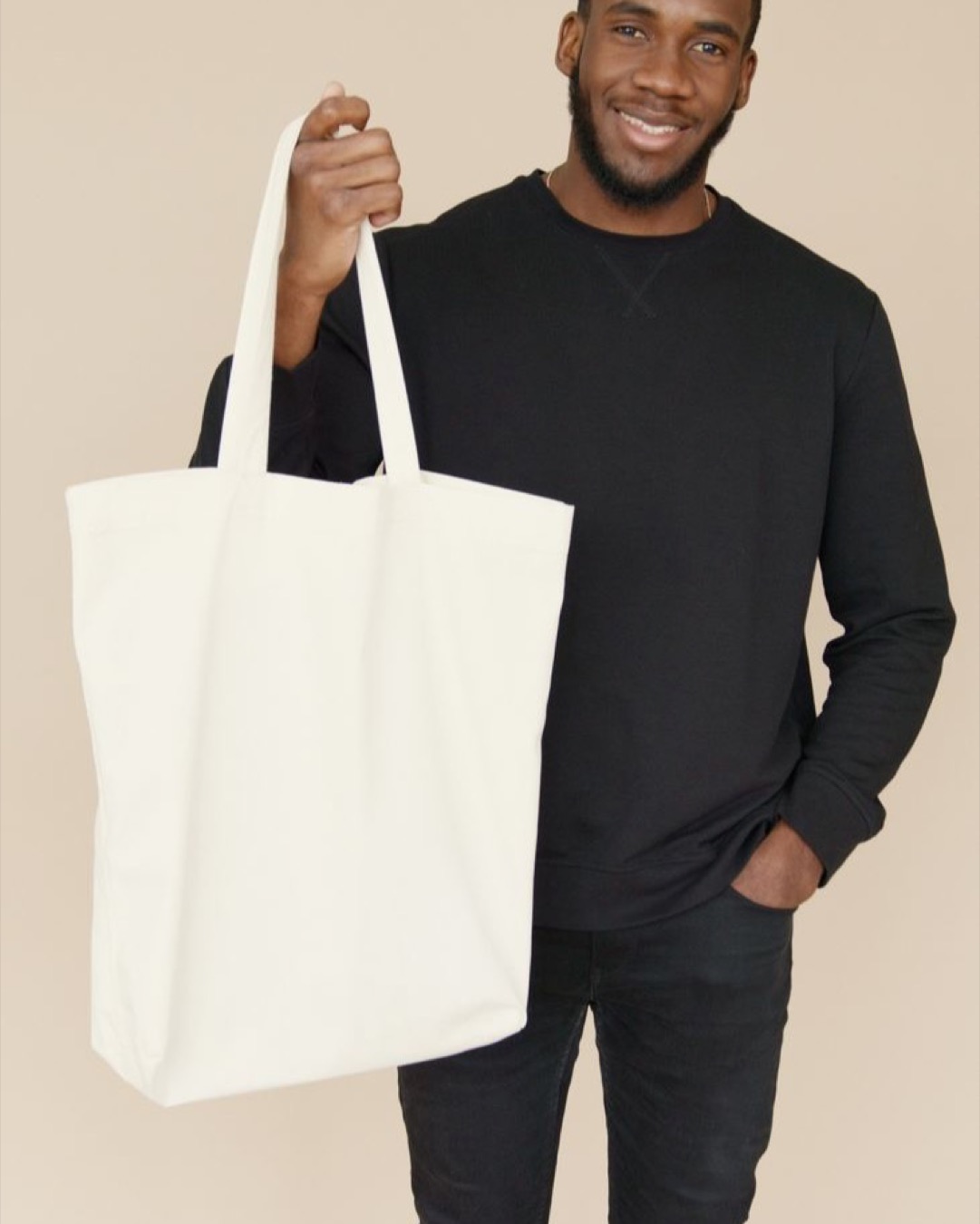 Smiling man in black attire showcasing a blank canvas tote bag against a neutral background.