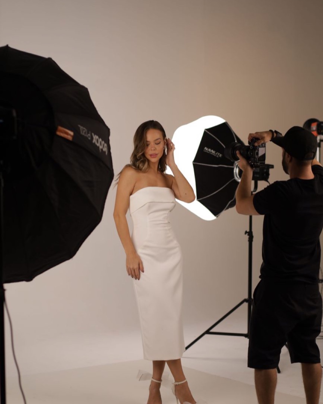 A model in a white dress poses during a professional studio photoshoot with lighting and camera setup.