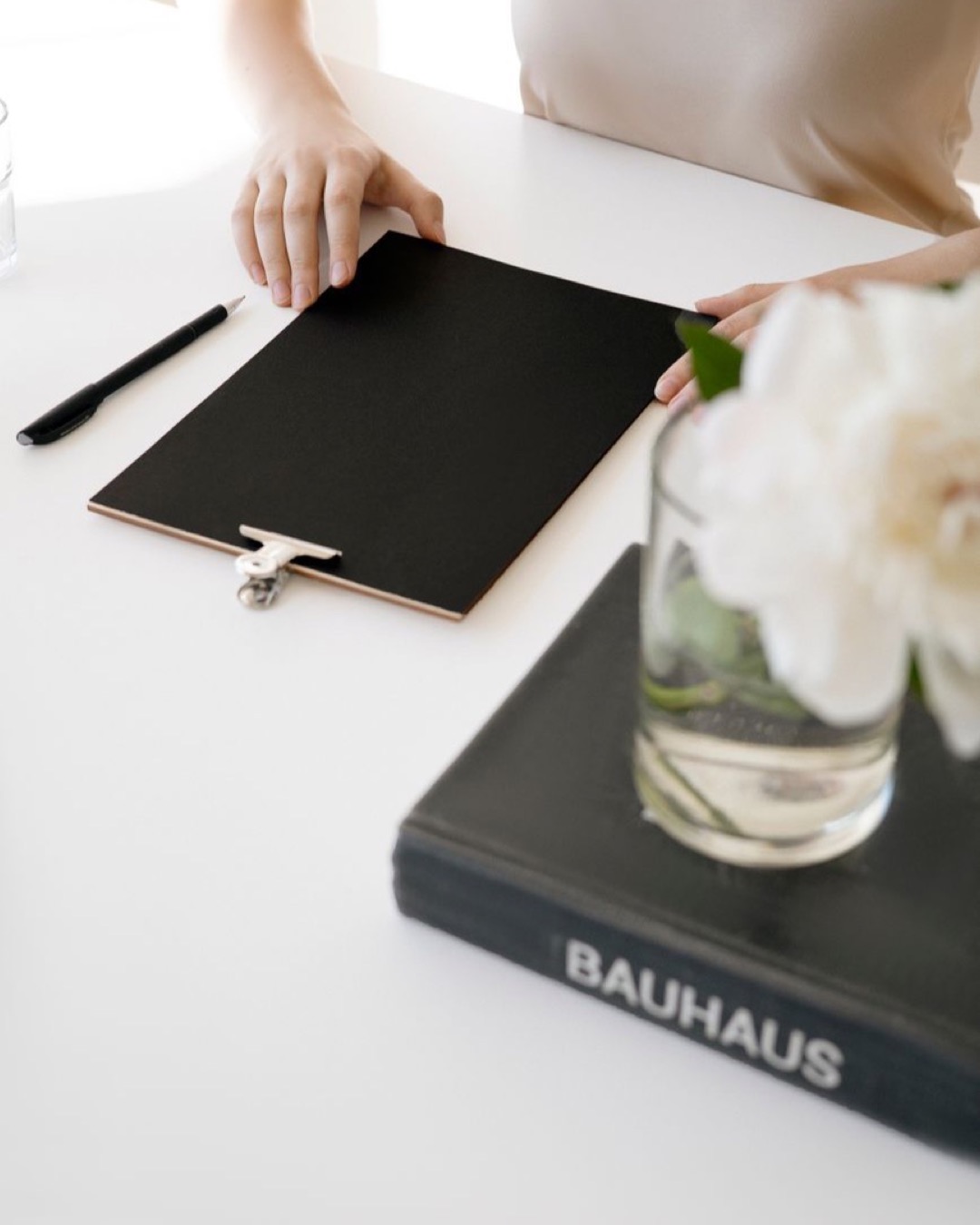 Aesthetic minimalist workspace featuring a clipboard, book, and flowers on a clean white desk.