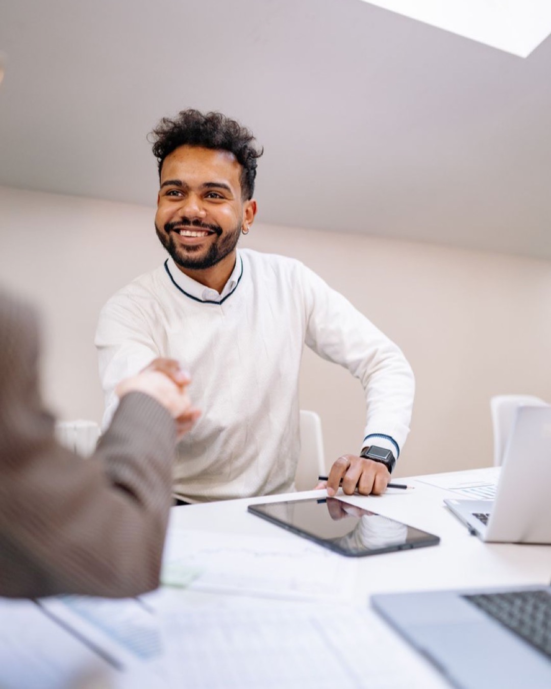 A smiling man in business attire shakes hands across a desk in a modern office.