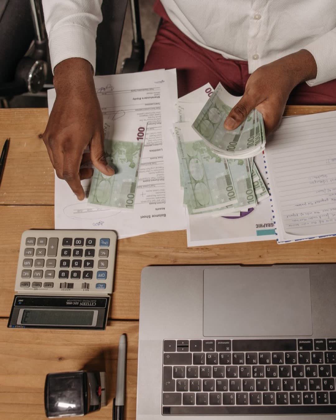 An overhead view of a professional counting cash and using a calculator to manage finances on a wooden desk.