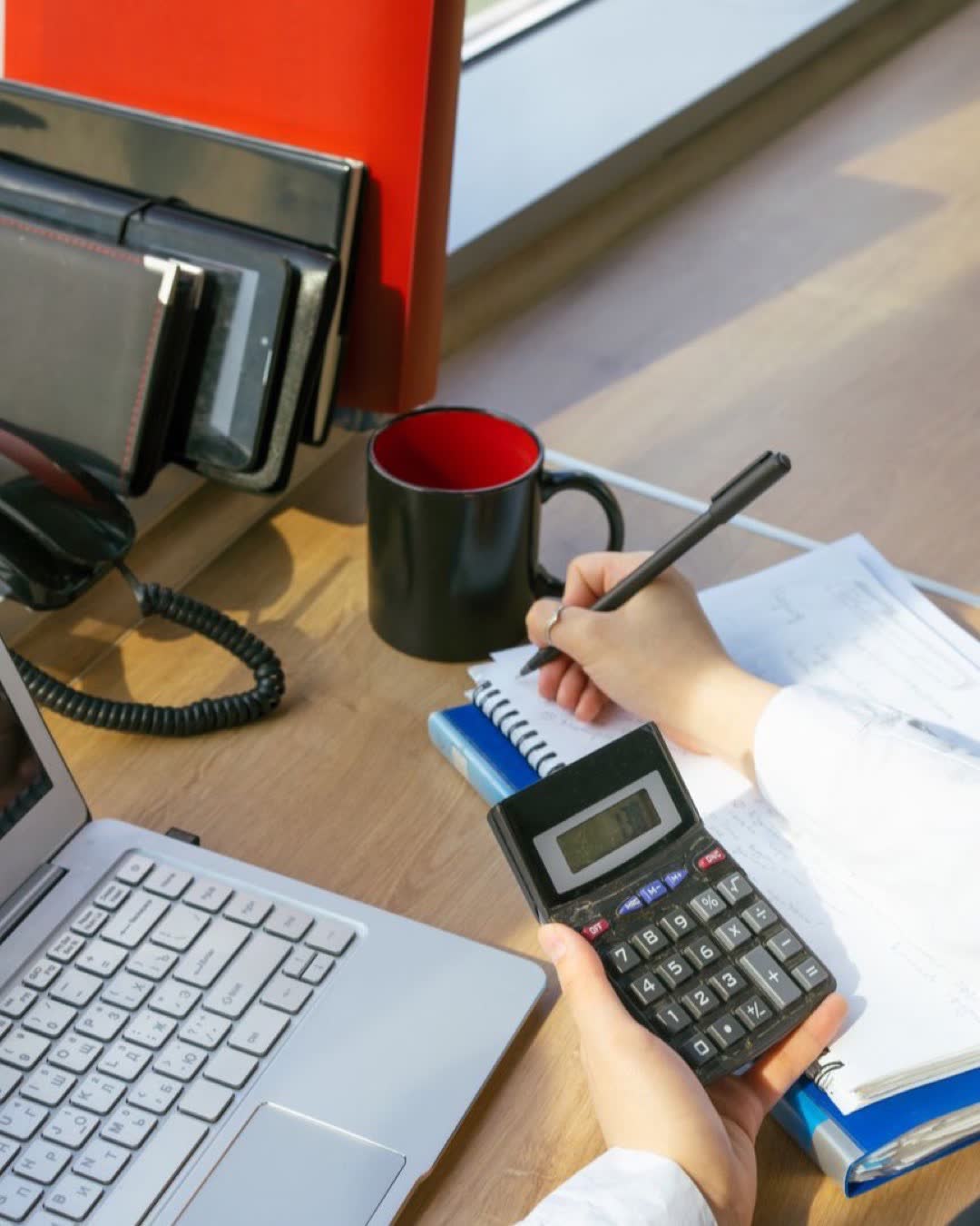 A person writing notes at a desk with a calculator, laptop, and red mug.