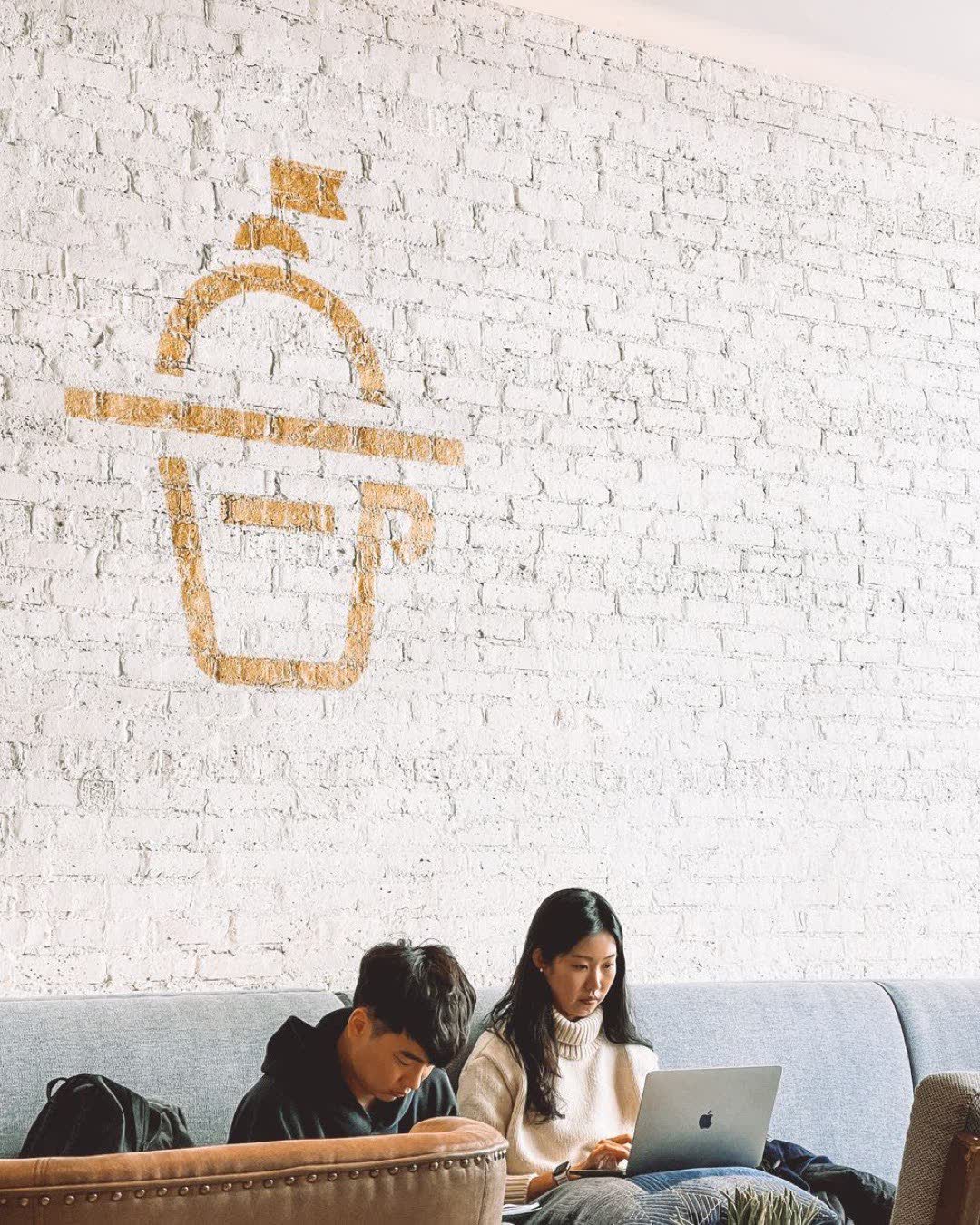 Two young adults focused on laptops, sitting in a chic cafe with a modern brick wall.