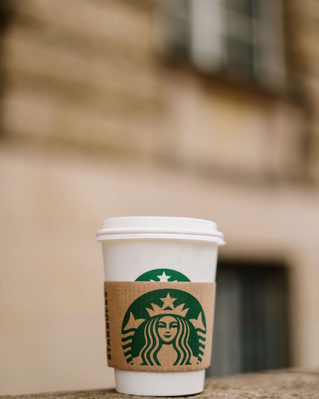A close-up shot of a coffee cup with a branded sleeve against a blurred outdoor background.