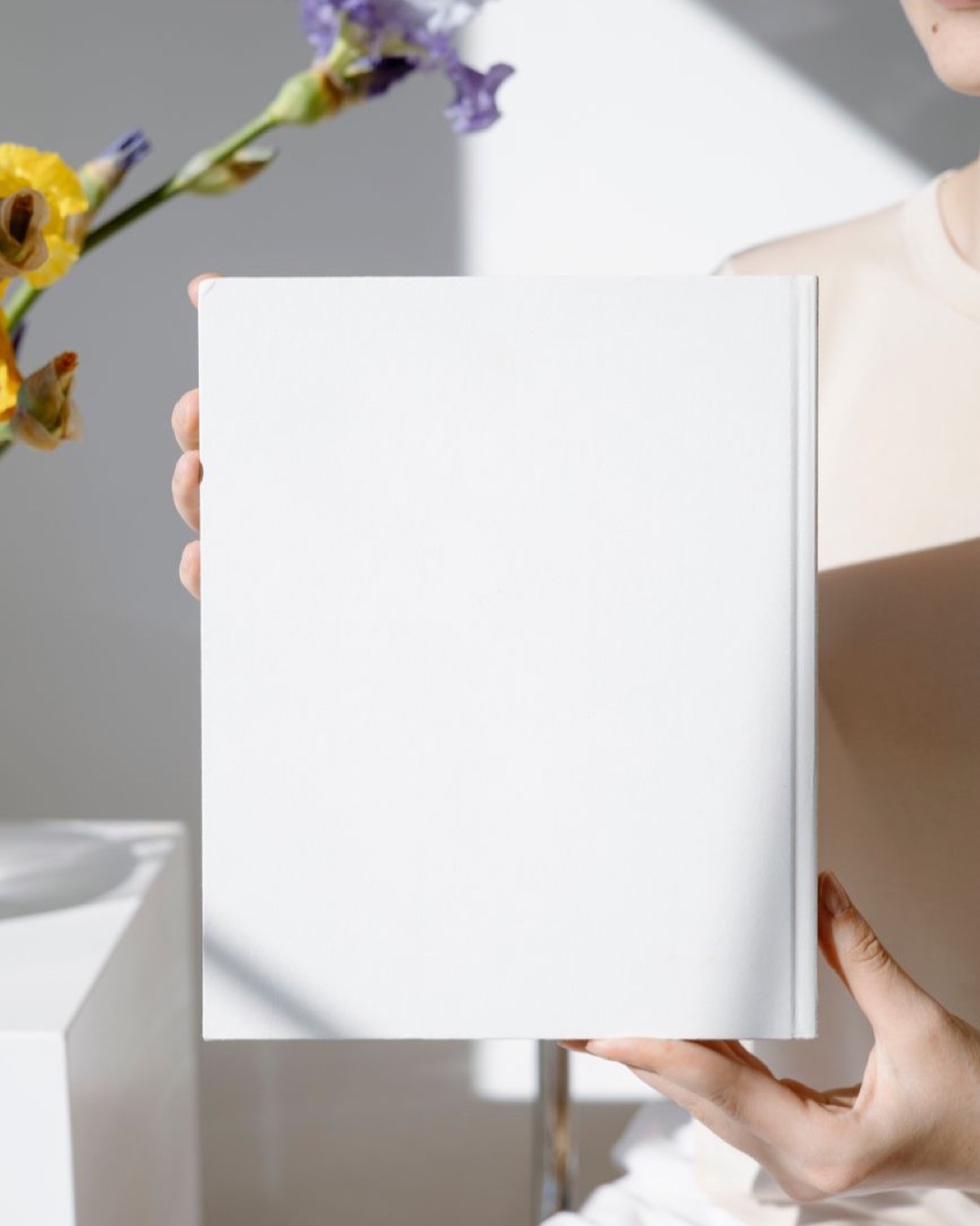 Minimalist blank book cover held in a bright room with flowers in the background.