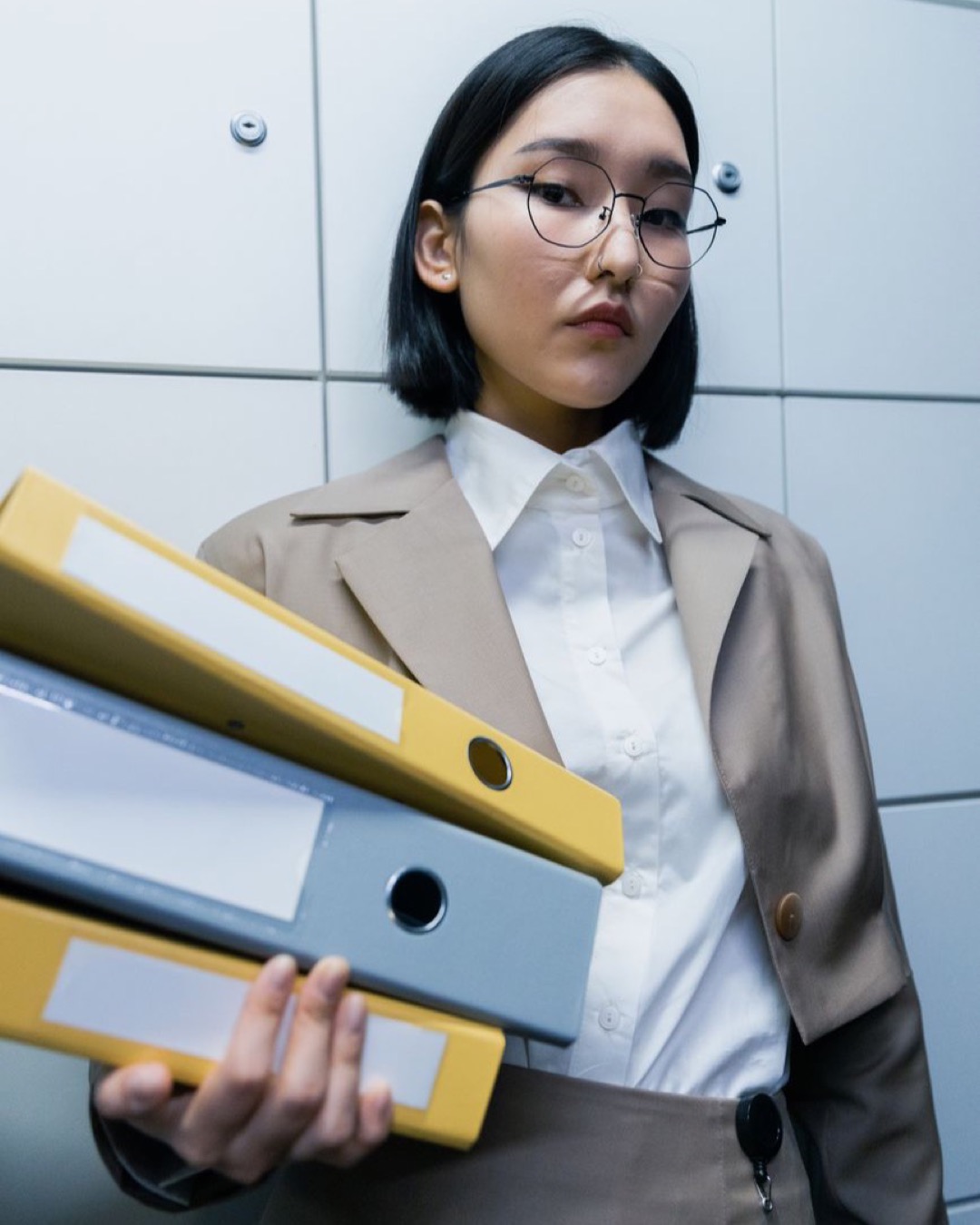 Asian businesswoman in a brown blazer holding binders in an office setting.