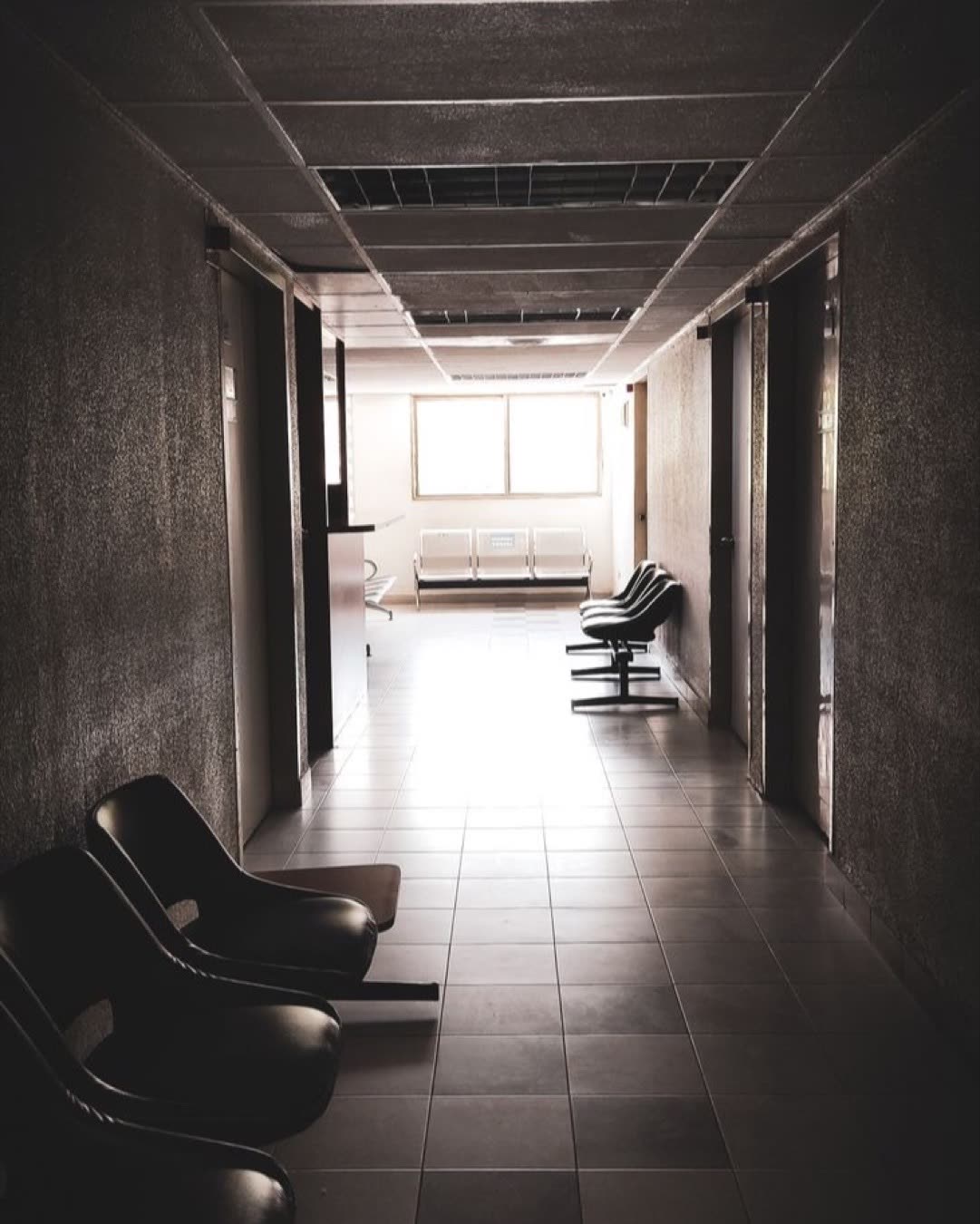 A dimly lit empty corridor with chairs and a waiting area, conveying a quiet atmosphere.