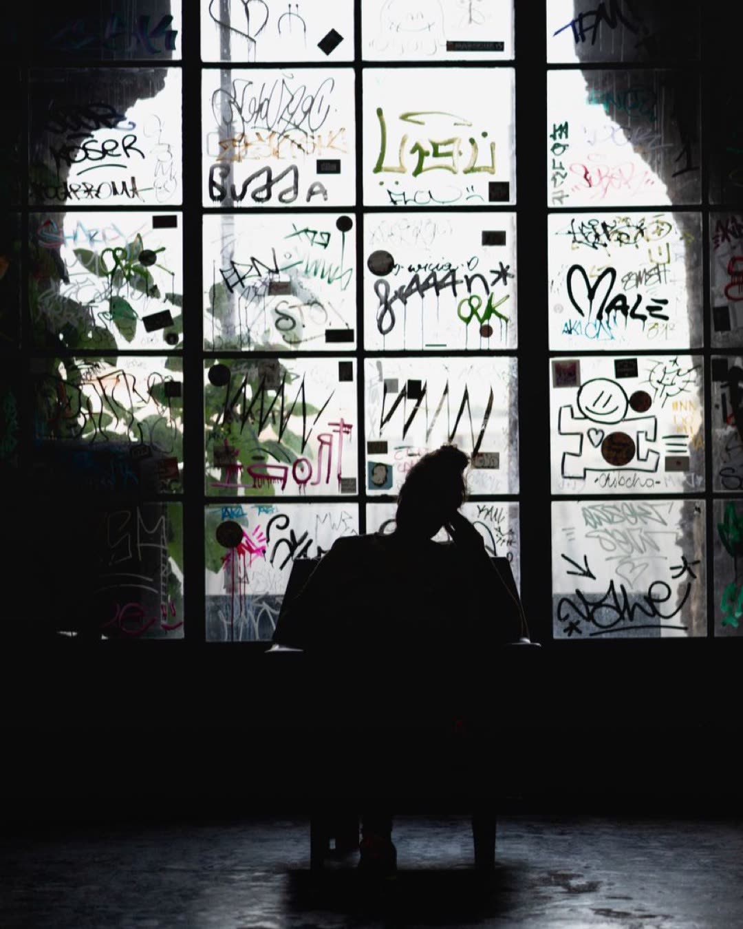 Silhouette of a person sitting thoughtfully against a window covered in graffiti art.