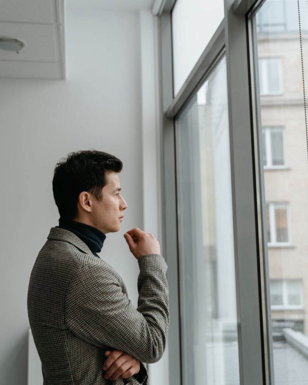 Side view of a thoughtful man in a coat looking out a large office window.