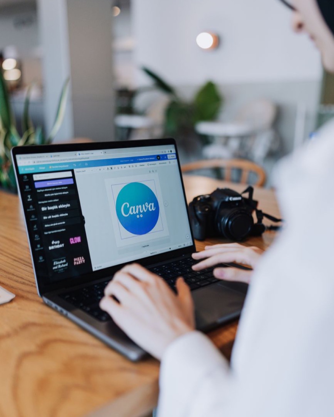 Young woman designing with Canva on laptop in a stylish indoor setting, accompanied by a digital camera.