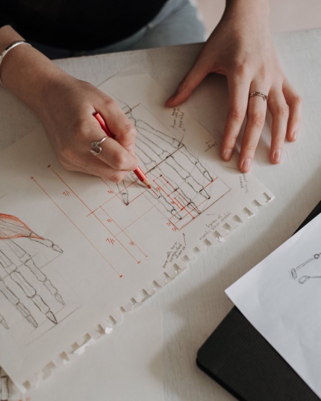 A detailed view of hands sketching anatomical drawings with red pencil on paper.