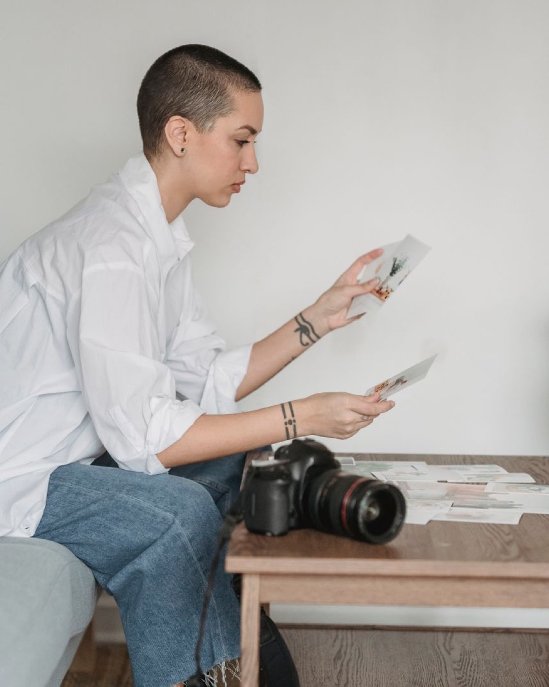 Side view of young female with tattoos comparing photos at desk with digital camera at home on light background