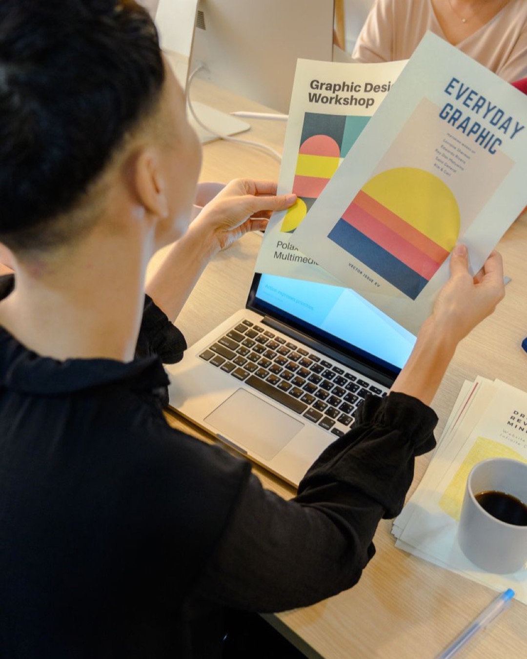 A professional reviews colorful graphic design brochures at a laptop in a modern office environment.