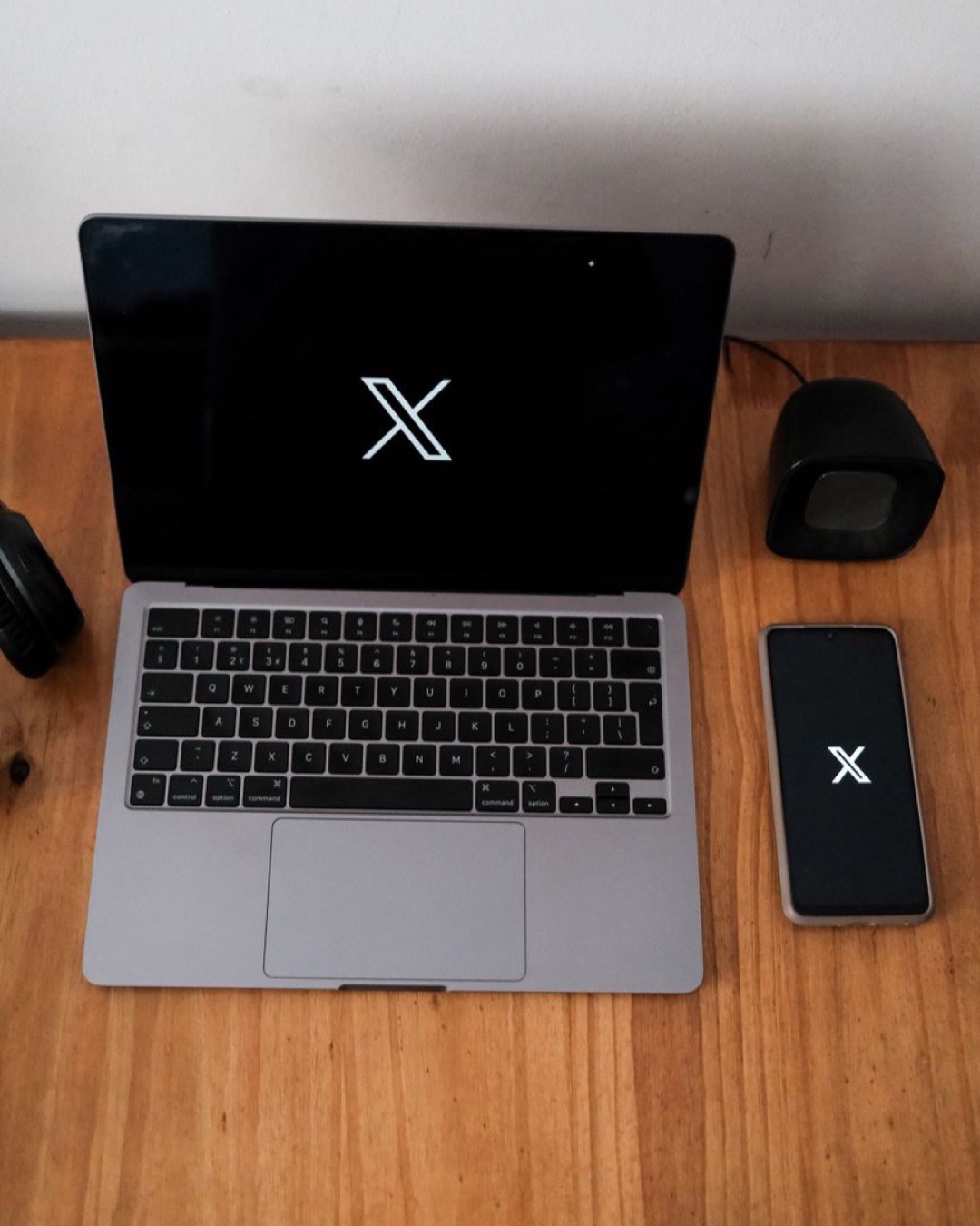 A minimalist setup featuring a laptop and smartphone displaying an X logo on wooden surface.