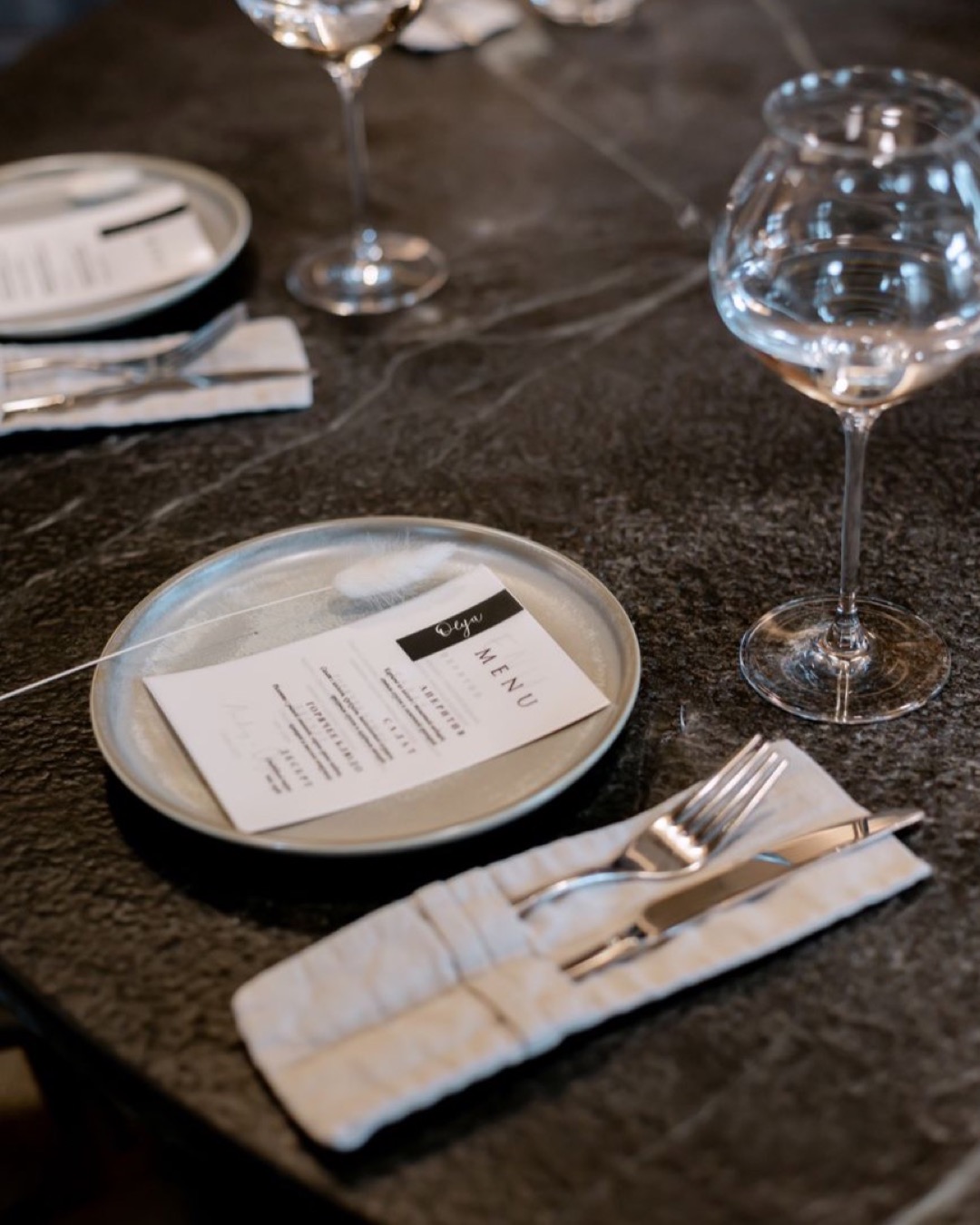 Stylish table setting in a restaurant with plates, cutlery, and menus on a dark textured surface.