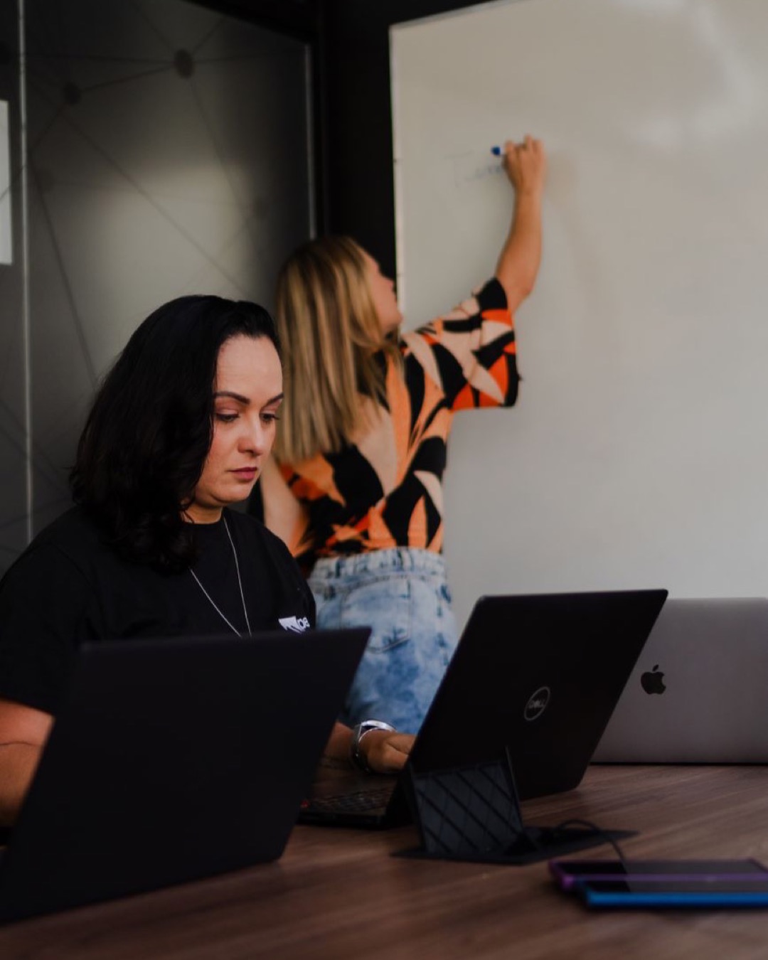Two women engaged in a collaborative office workspace, one writing on a whiteboard.