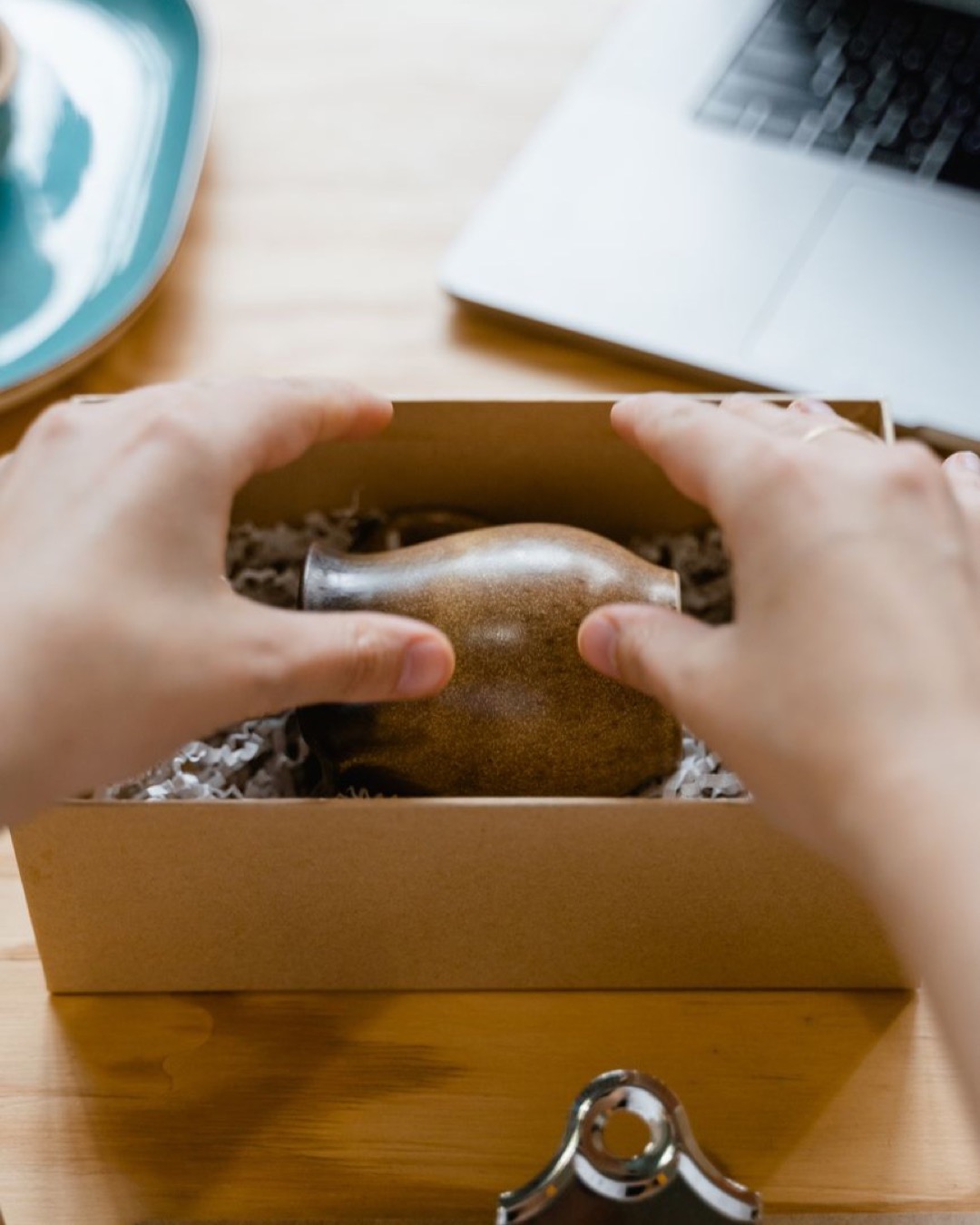 Person packaging a ceramic pot into a cardboard box on a wooden table.