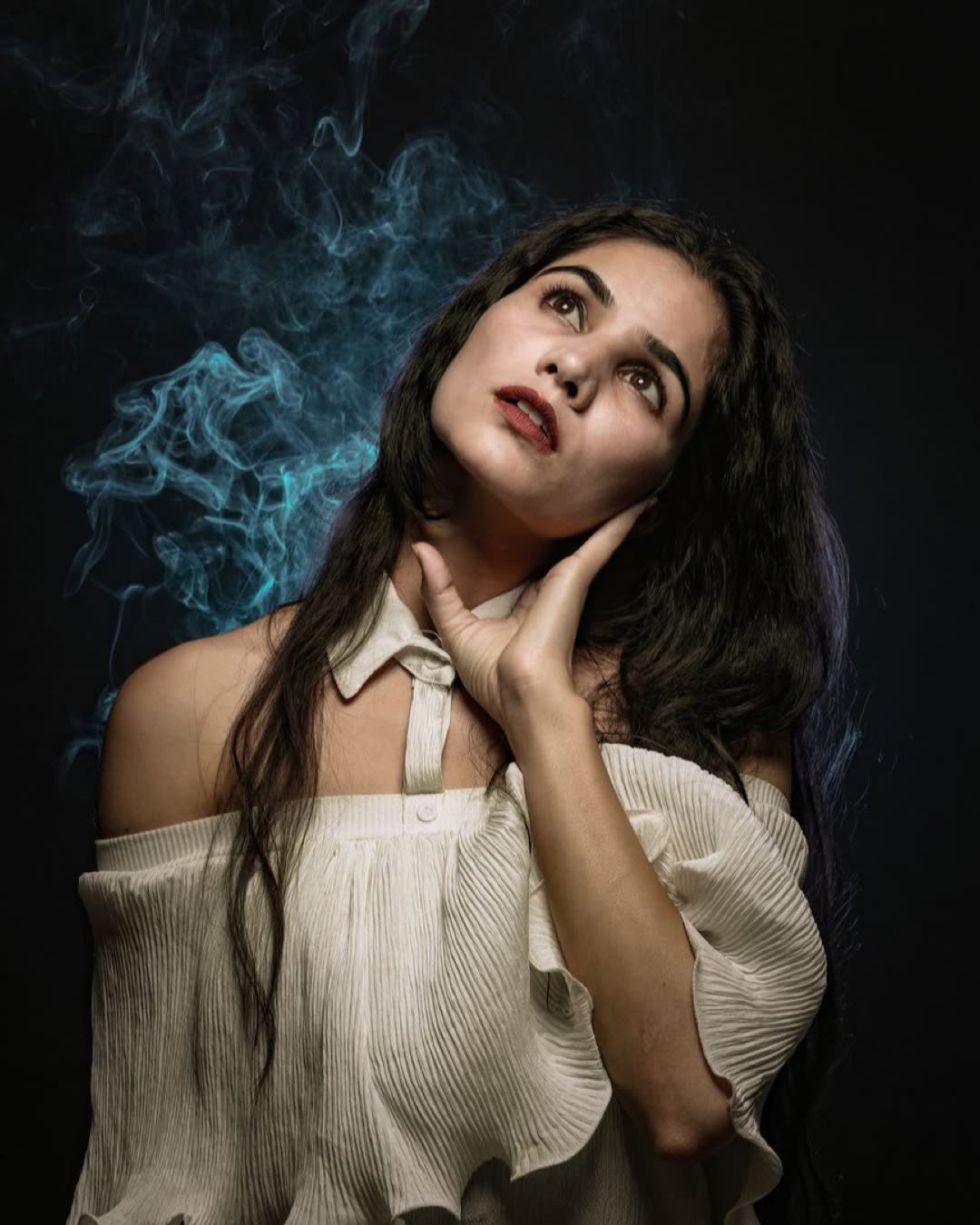 Beautiful serene brunette in stylish white outfit with bare shoulders touching face tenderly and looking up while standi