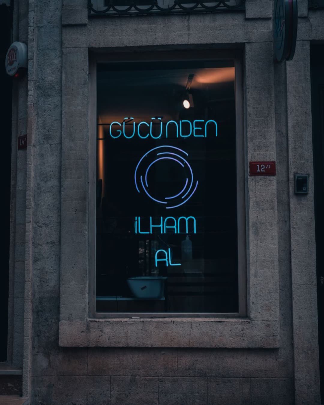Storefront with neon sign reading 'Güçünden İlham Al' in a moody urban street setting.