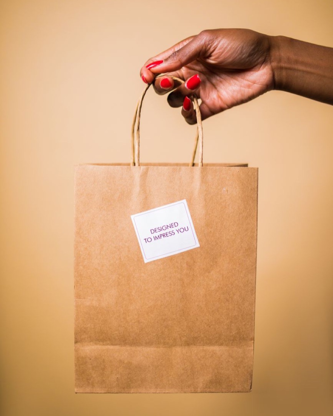 A hand holding a paper bag with a note reading 'Designed to Impress You' against a beige background.
