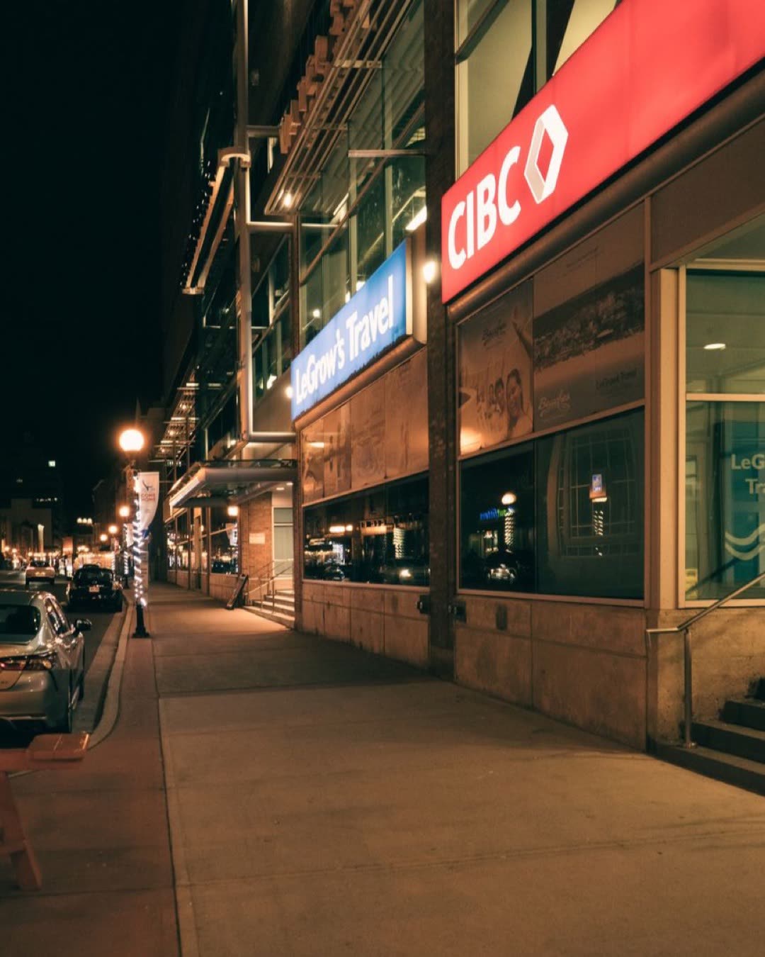 City street scene with illuminated signs including CIBC and LeGrow's Travel at night.
