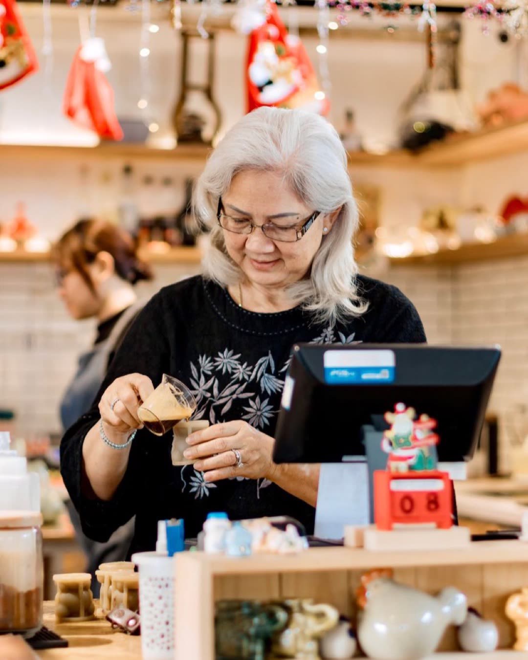 Elderly woman in a café prepping a drink during holiday season. Cozy and festive.