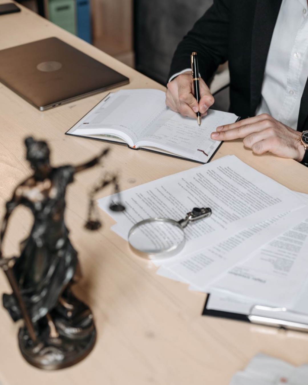 Professional working on documents at desk with legal statue and magnifying glass.