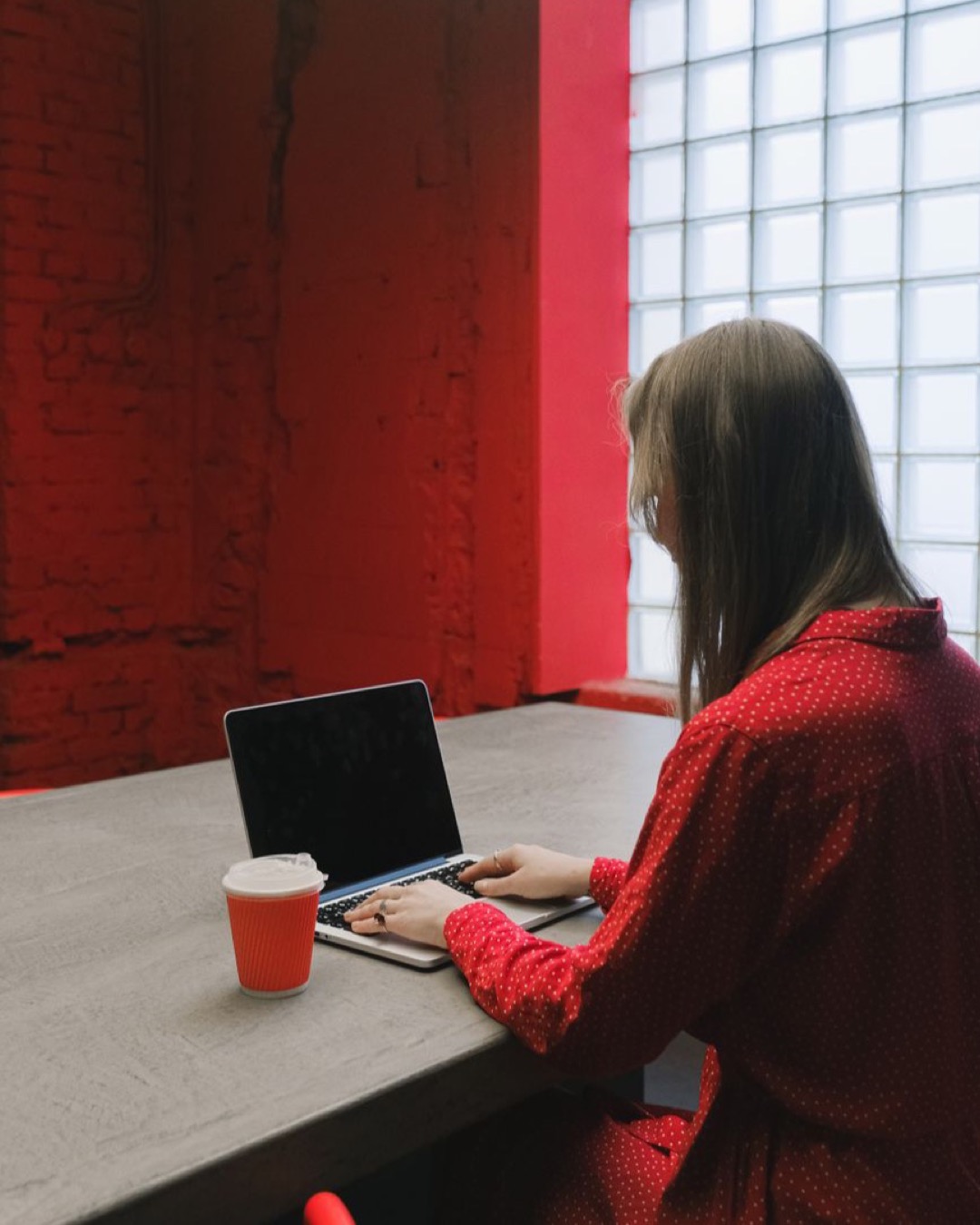 Woman in a red dress working on a laptop at a desk with a coffee cup indoors.
