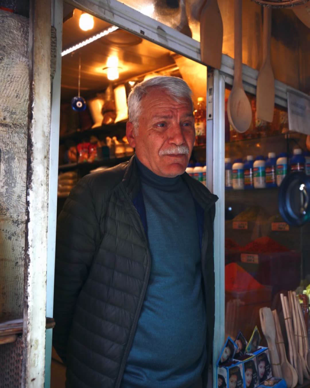 Senior man with a mustache stands at shop entrance, surrounded by goods.